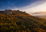Herbstlandschaft mit bewaldeten Hügeln unter blauem Himmel und einem Felsen im Hintergrund.