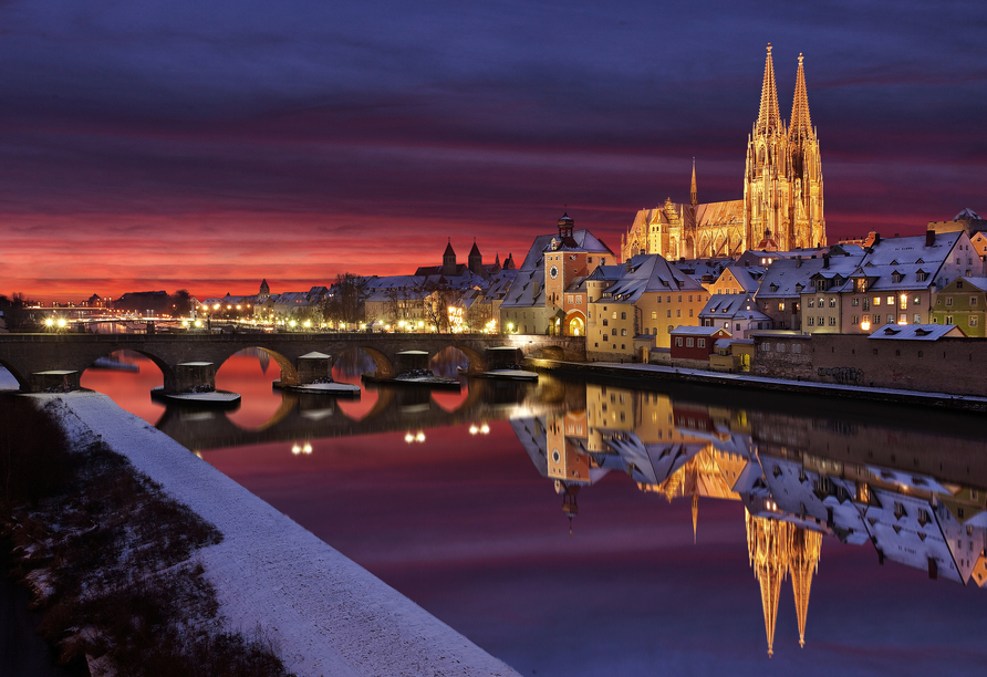 Blick auf die verschneite Altstadt von Regensburg in Bayern mit der Steinernen Brücke und dem Dom St. Peter bei Sonnenuntergang im Winter