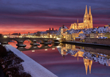 Blick auf die verschneite Altstadt von Regensburg in Bayern mit der Steinernen Brücke und dem Dom St. Peter bei Sonnenuntergang im Winter