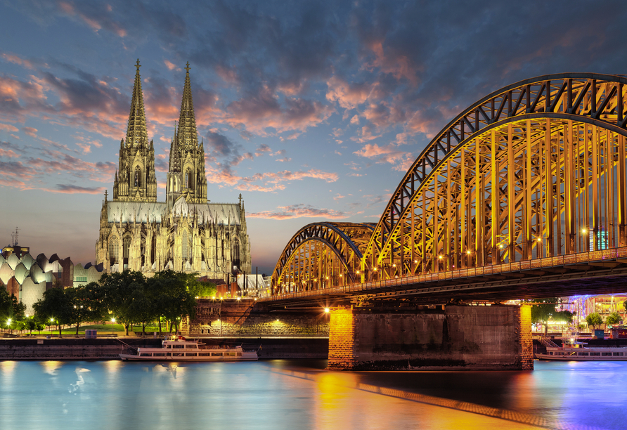 Der Kölner Dom und die Lichter der Stadt spiegeln sich im Rhein zur Dämmerung.