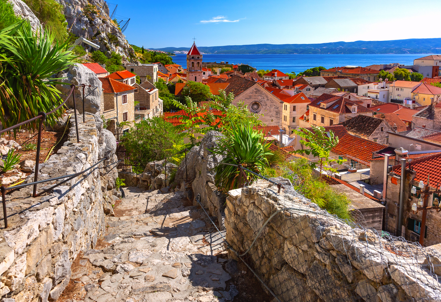 Ein malerischer Pfad führt durch die Altstadt von Omiš, vorbei an alten Steinmauern und roten Dächern, bis zum funkelnden Blau der Adria, das in der Sonne glitzert.