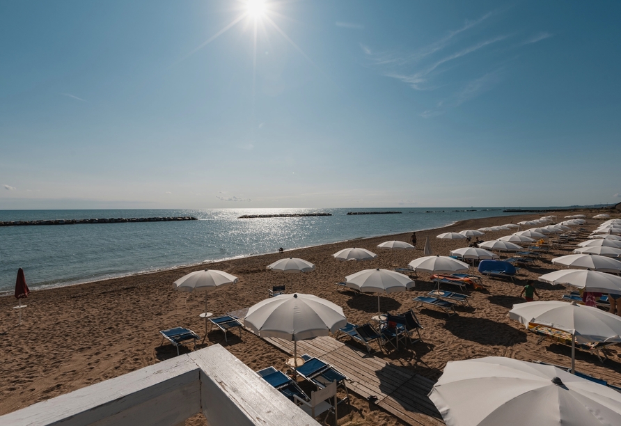 Sonniger Strand mit Liegen und Schirmen direkt am glitzernden Meer